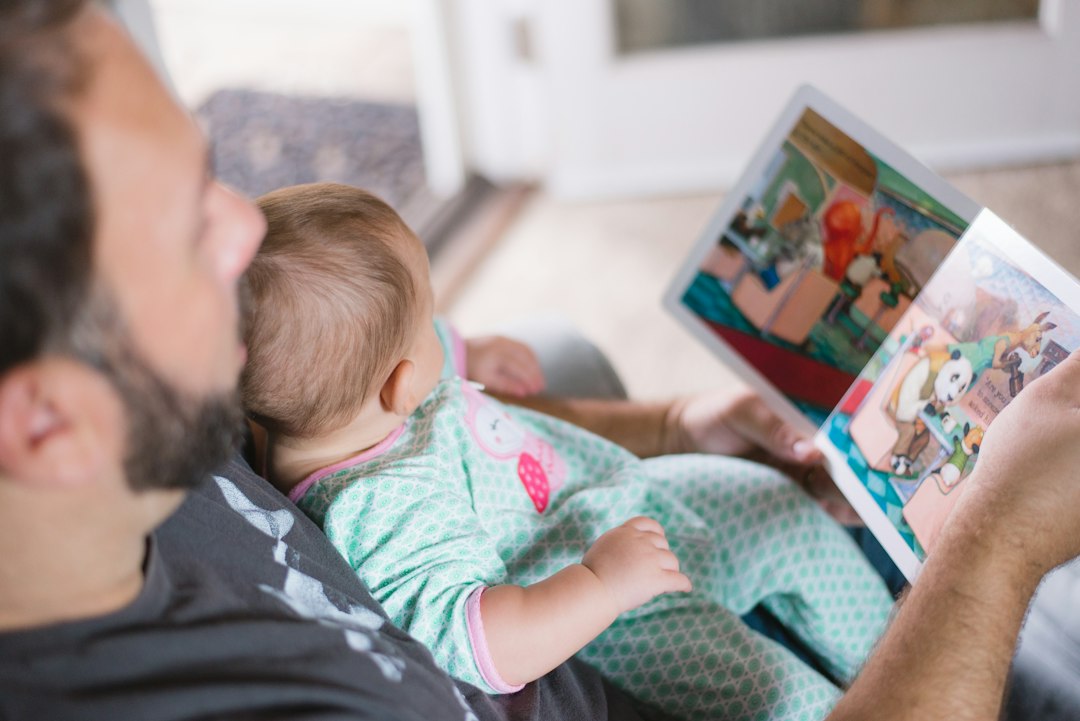 Egy apa az ölében tartott kisbabájának képeskönyvet mutat otthon.