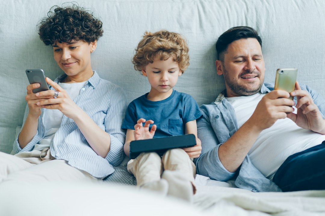 Egy család a kanapén ül, a szülők a telefonjukat, a gyermek egy tabletet néz.