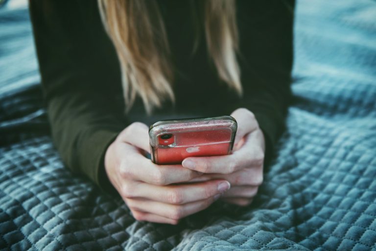 Egy kamasz kezében telefon, az ágyon fekve, arca nem látszik.