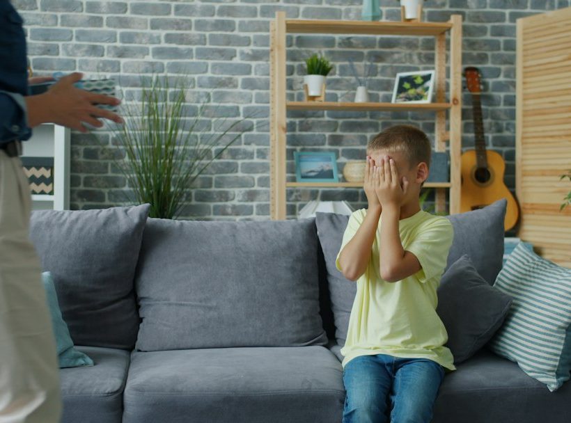 Kisfiú a kanapén eltakarja az arcát, előtte egy felnőtt áll.