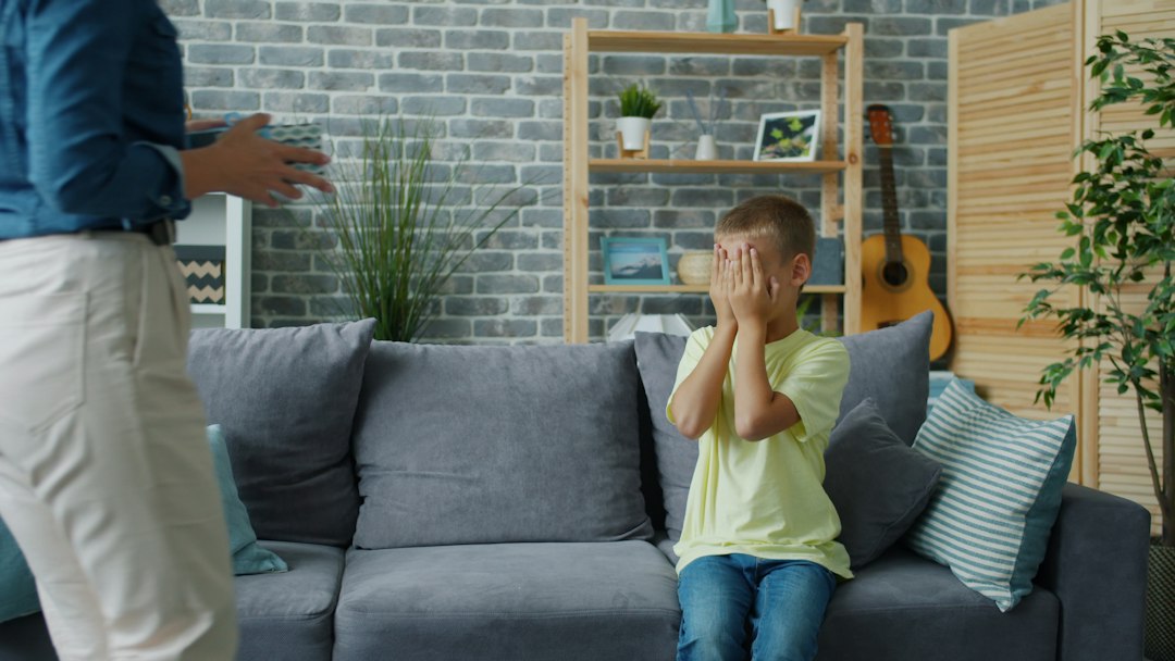 Kisfiú a kanapén eltakarja az arcát, előtte egy felnőtt áll.