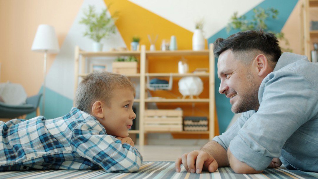 Egy apa és kisfia a földön fekve, mosolyogva beszélgetnek otthon.