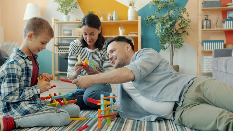 Szülők és kisfiuk együtt építenek színes építőelemekből a nappali szőnyegén.