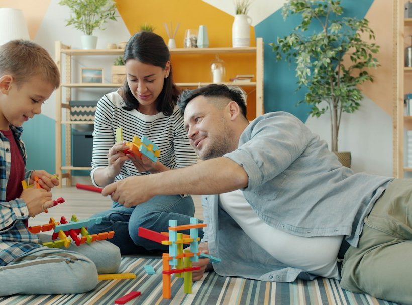 Szülők és kisfiuk együtt építenek színes építőelemekből a nappali szőnyegén.