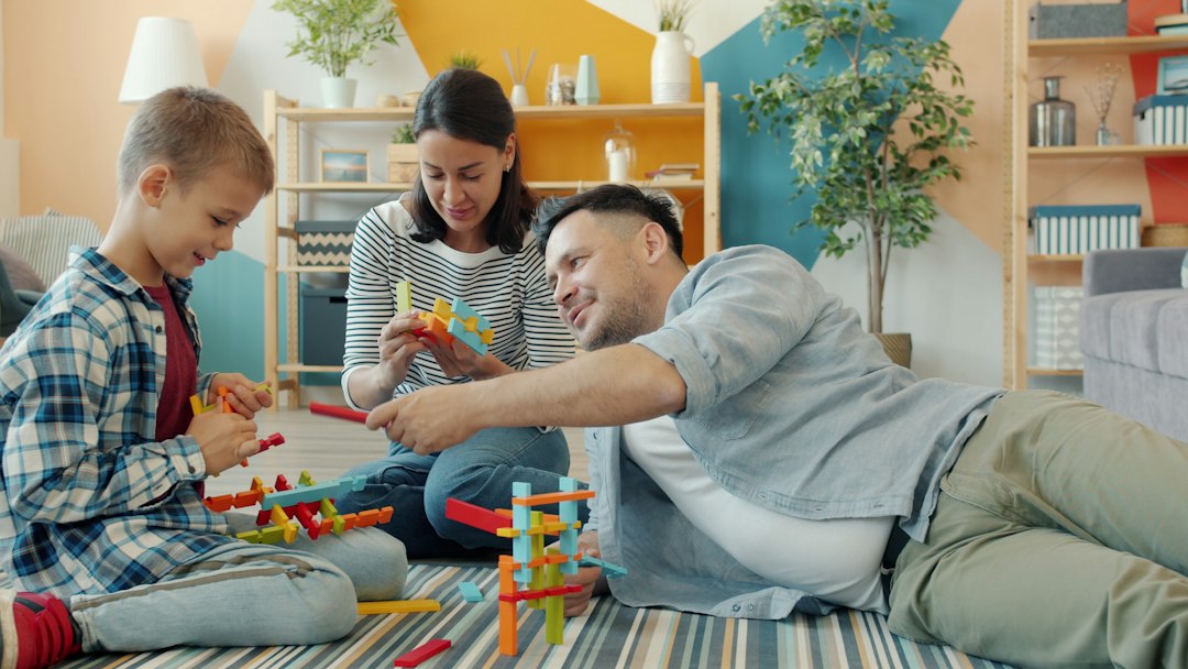 Szülők és kisfiuk együtt építenek színes építőelemekből a nappali szőnyegén.