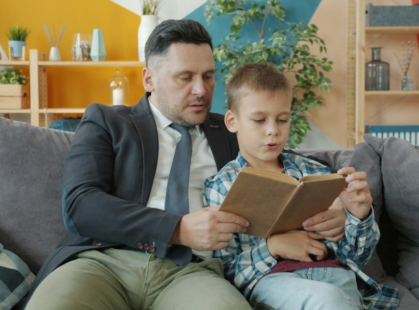 Apa és kisfia a kanapén ülve együtt olvas egy könyvet.